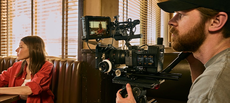 Εικόνα χρήσης μιας φωτογραφικής μηχανής FX5 σε εξάρτημα λήψης για τον ώμο ενός χρήστη που κινηματογραφεί ένα άτομο που κάθεται σε μια καφετέρια, φωτισμένο από φυσικό φως από τα παράθυρα.